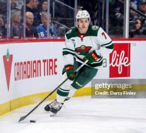 Ice hockey player in a white and green jersey (number 7) skating along the boards with a stick and puck during a game, with spectators in the stands in the background.
