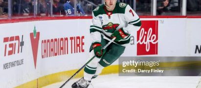 Ice hockey player in a white and green jersey (number 7) skating along the boards with a stick and puck during a game, with spectators in the stands in the background.