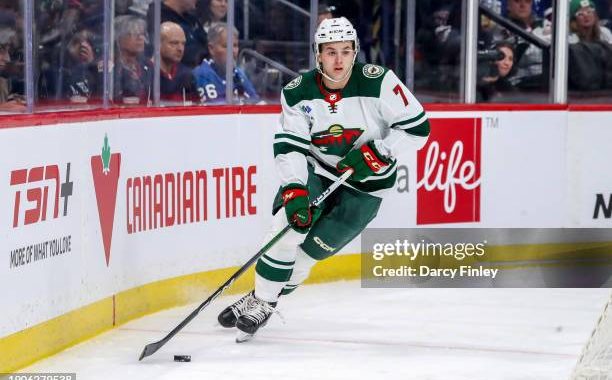 Ice hockey player in a white and green jersey (number 7) skating along the boards with a stick and puck during a game, with spectators in the stands in the background.