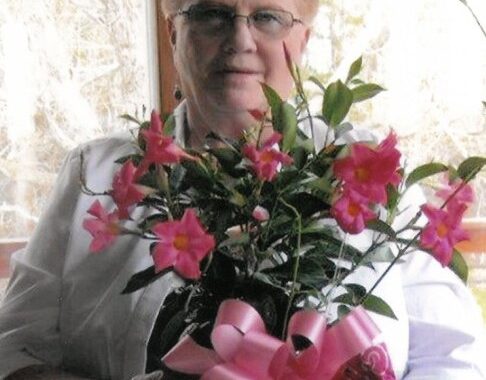 Older woman with short light hair and glasses holding a pink-flowered bouquet with a pink ribbon bow.