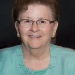 Portrait of an elderly woman with short brown hair and glasses, wearing a turquoise cardigan and smiling at the camera.