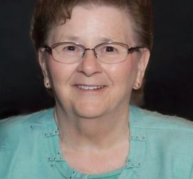 Portrait of an elderly woman with short brown hair and glasses, wearing a turquoise cardigan and smiling at the camera.