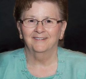 Portrait of an elderly woman with short brown hair and glasses, wearing a turquoise cardigan and smiling at the camera.