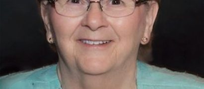 Portrait of an elderly woman with short brown hair and glasses, wearing a turquoise cardigan and smiling at the camera.