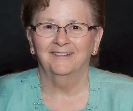 Portrait of an elderly woman with short brown hair and glasses, wearing a turquoise cardigan and smiling at the camera.