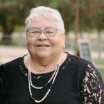 Smiling elderly woman with short white hair and blue glasses, wearing a black lace dress and pearl necklace outdoors.