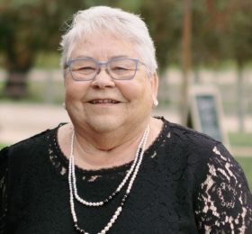 Smiling elderly woman with short white hair and blue glasses, wearing a black lace dress and pearl necklace outdoors.