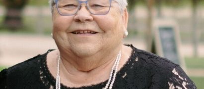 Smiling elderly woman with short white hair and blue glasses, wearing a black lace dress and pearl necklace outdoors.
