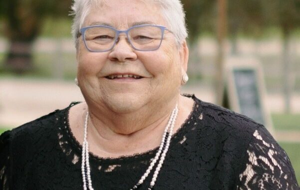 Smiling elderly woman with short white hair and blue glasses, wearing a black lace dress and pearl necklace outdoors.