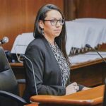 Professional woman in a black blazer and glasses sits at a wooden podium in a courtroom, speaking into a microphone.