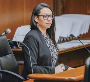 Professional woman in a black blazer and glasses sits at a wooden podium in a courtroom, speaking into a microphone.