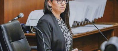 Professional woman in a black blazer and glasses sits at a wooden podium in a courtroom, speaking into a microphone.