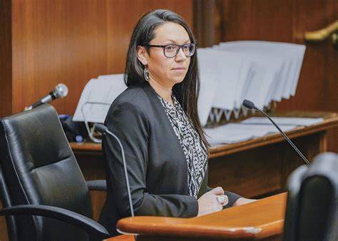 Professional woman in a black blazer and glasses sits at a wooden podium in a courtroom, speaking into a microphone.